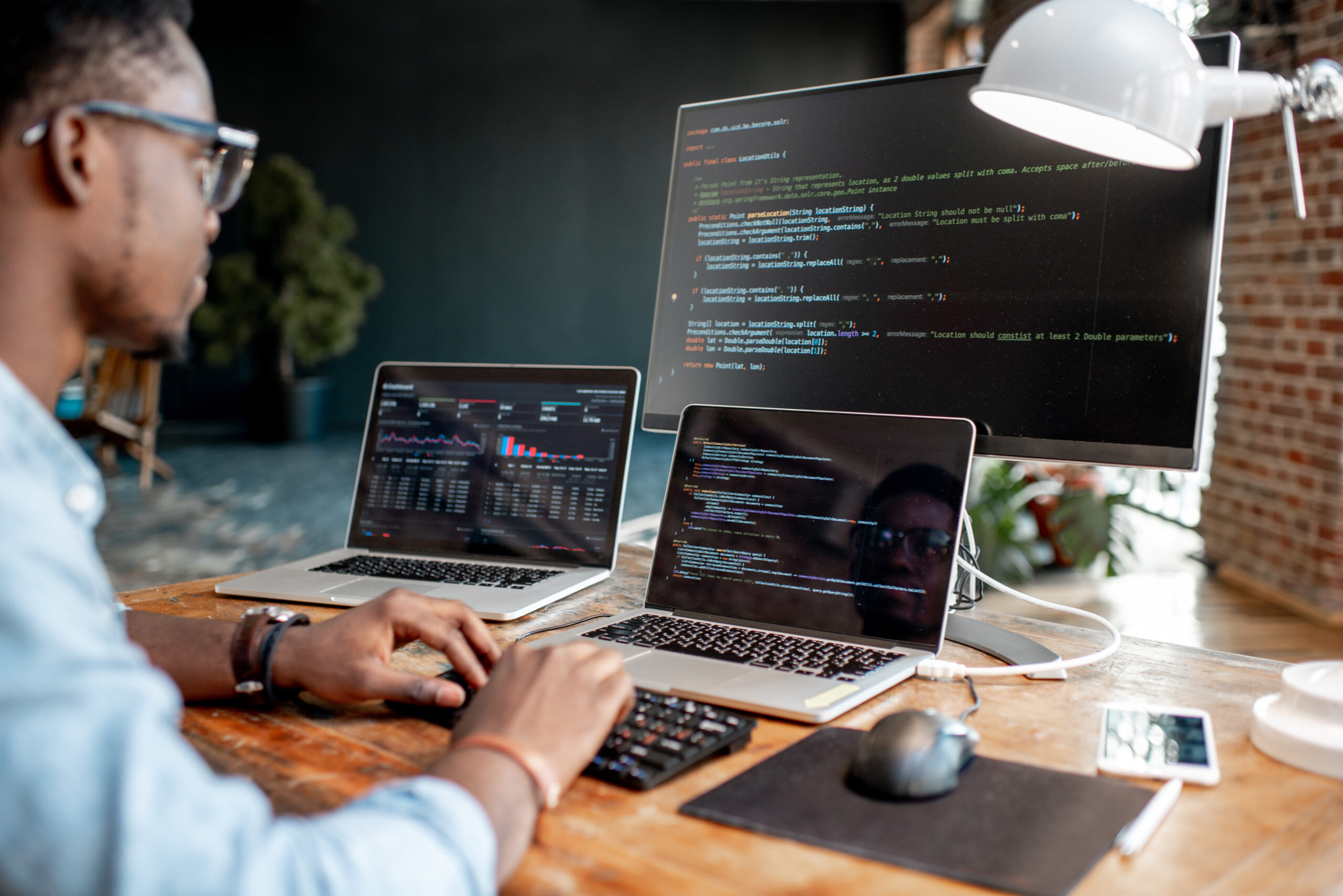 Young african male programmer writing program code sitting at the workplace with three monitors in the office. Image focused on the screen